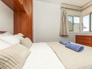 A neatly made bed with a rolled towel, a wooden headboard, a bedside table with a lamp, and a window with curtains.
