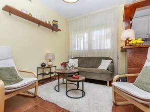 A living room with a sofa, two armchairs, a coffee table, a lamp, a small table with a bowl of fruit, and a shelf with decorative items.