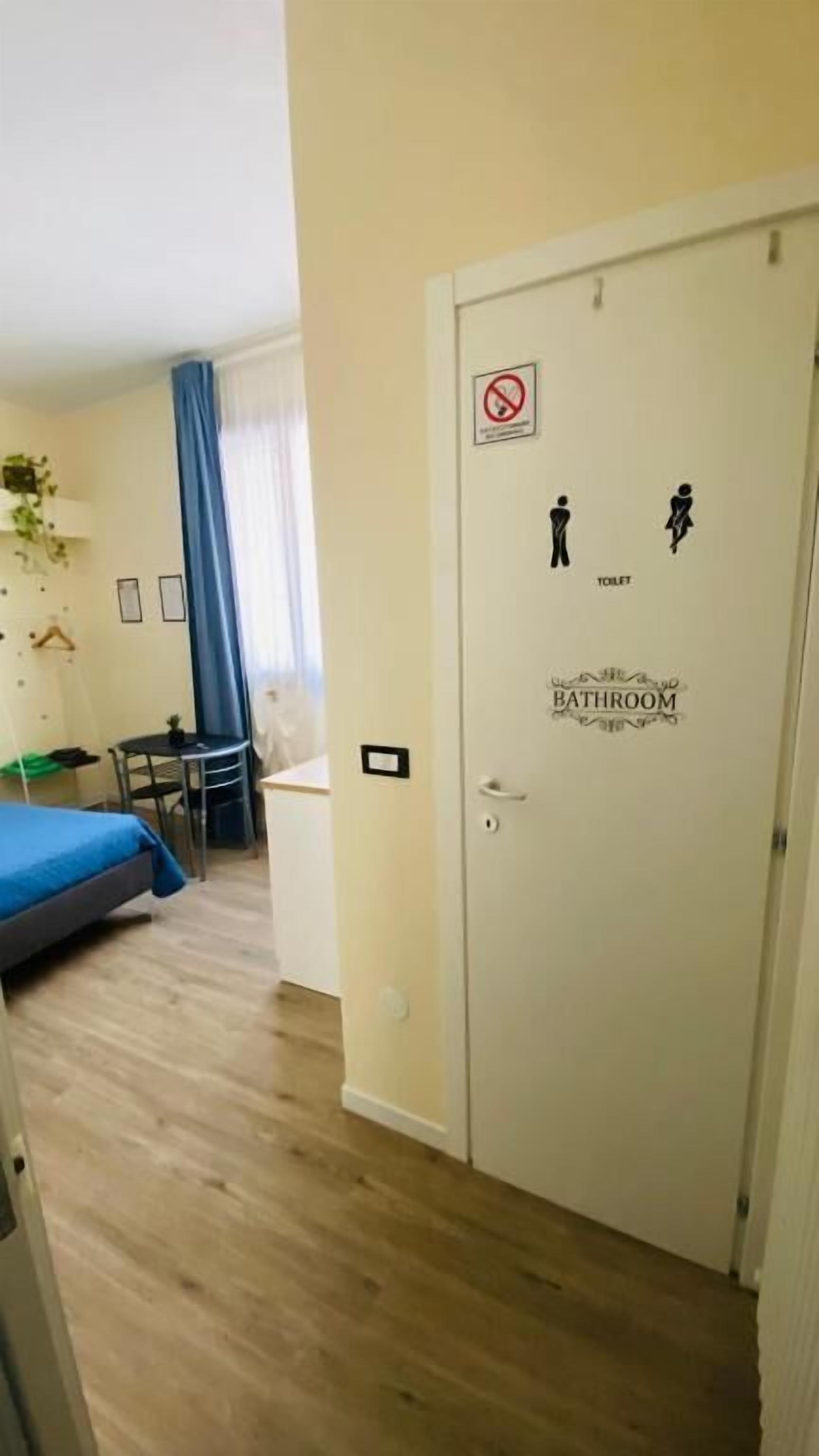 A hotel room with a bed, a small table, a chair, a plant, and a door with a bathroom sign.