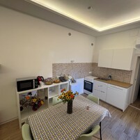 Cocina compacta con microondas, hervidor y horno pequeño. Hay una mesa de comedor con mantel estampado y un florero con flores.
