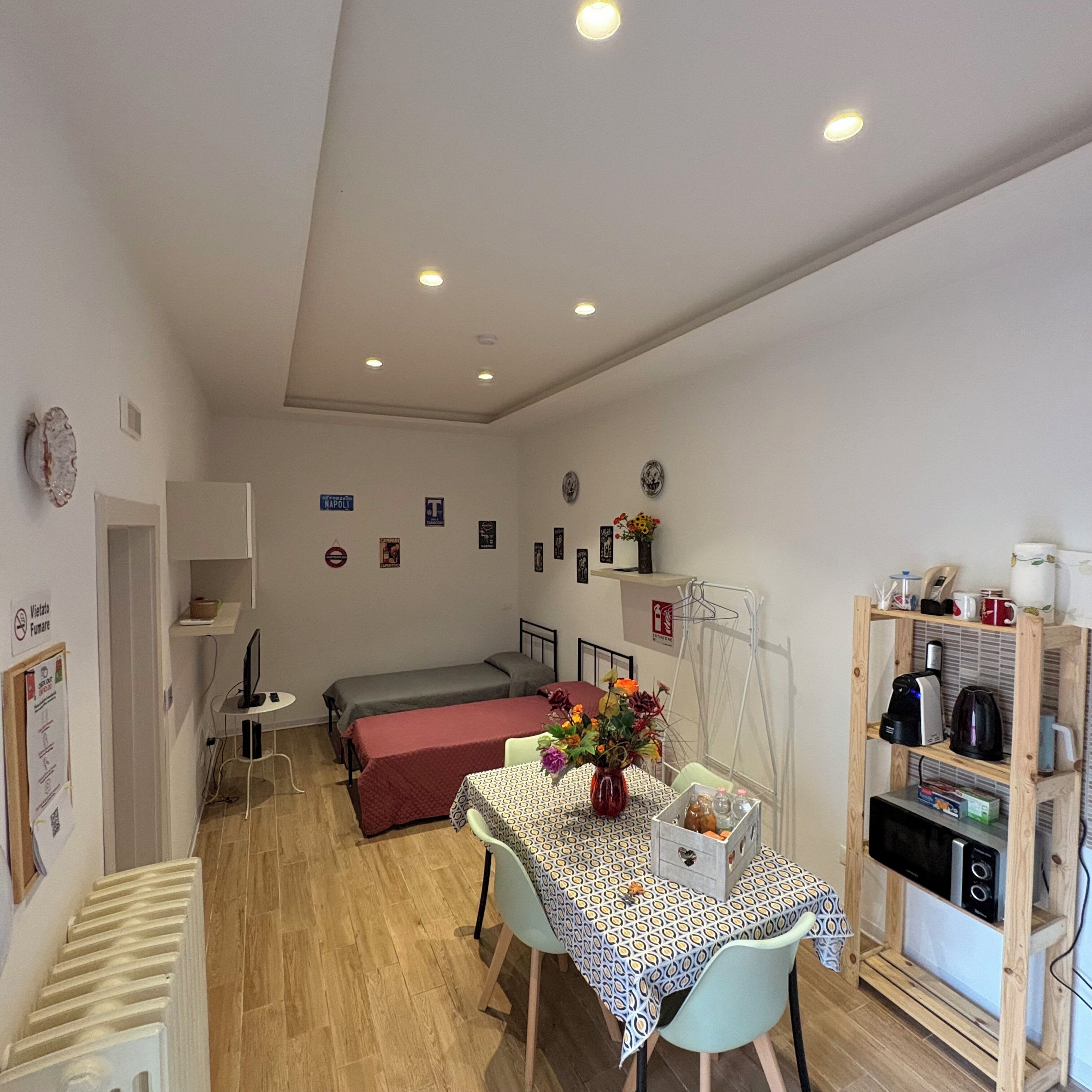 Una habitación pequeña y bien iluminada con una cama, una mesa con un florero y una zona de kitchenette.