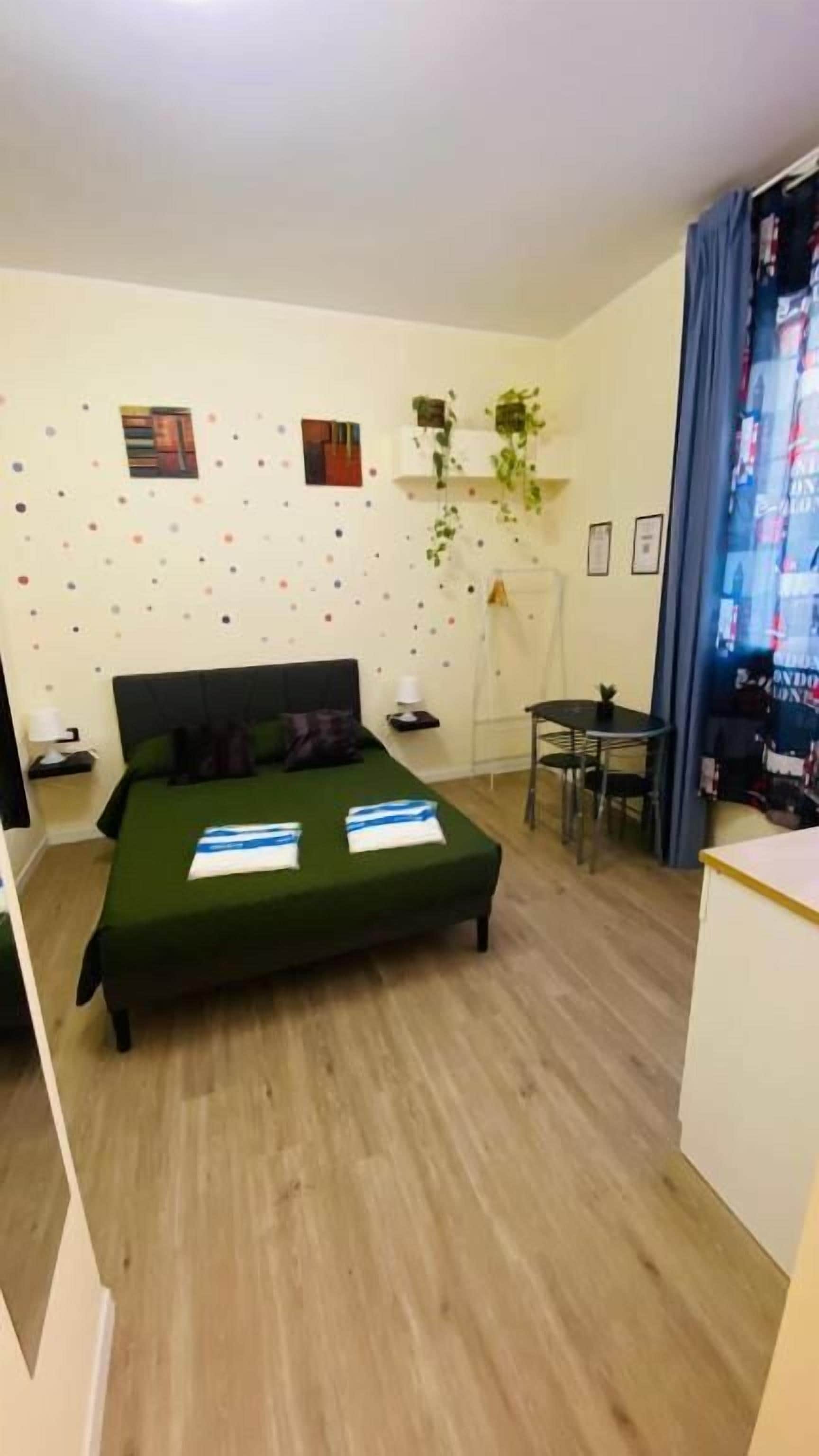 A small, clean room with a bed, a desk, and a chair. There are wall decorations and a window with curtains.