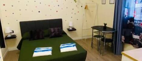 A small, clean room with a bed, a desk, and a chair. There are wall decorations and a window with curtains.