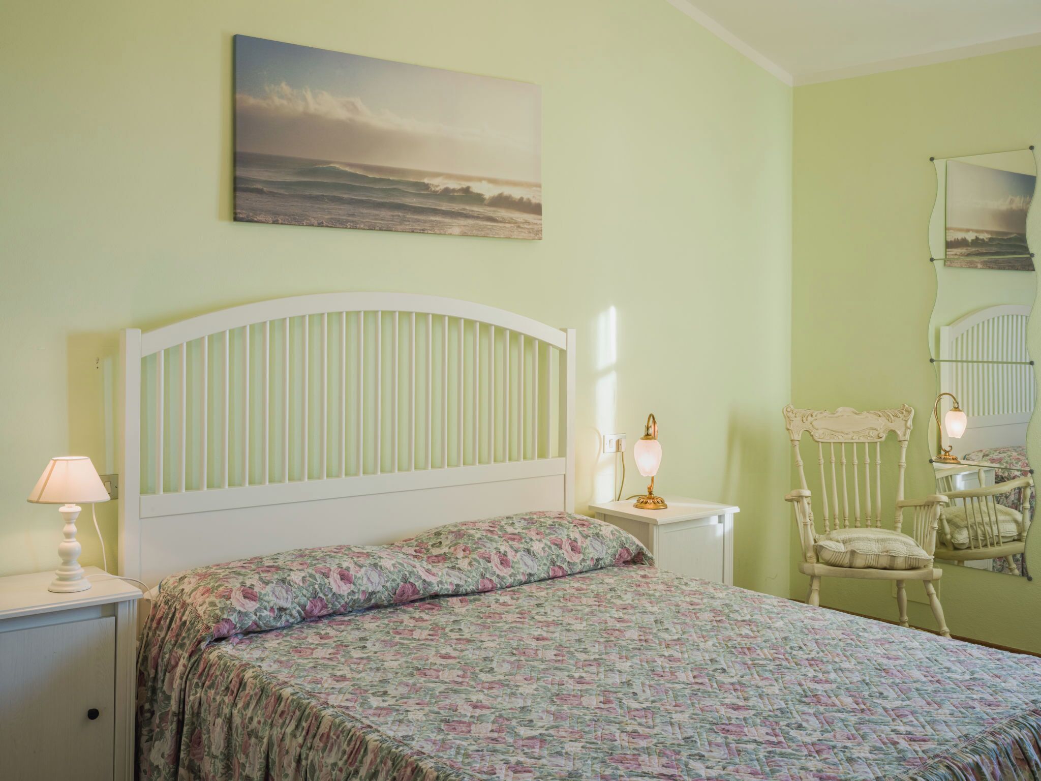 Un dormitorio con cama, mesita de noche, silla, lámpara y un cuadro en la pared.