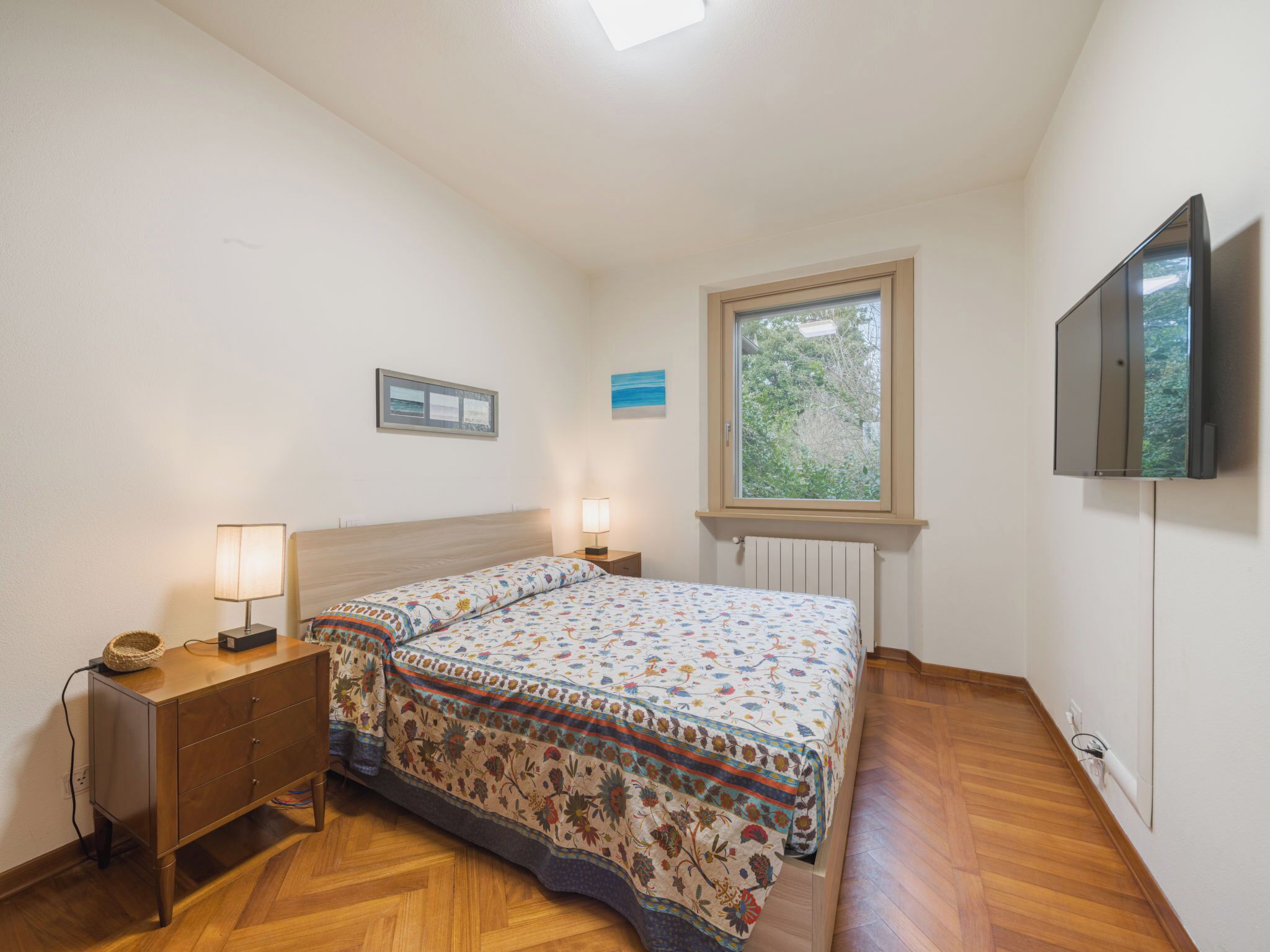 A bedroom with a bed, bedside lamps, a dresser, a window with a view, and a TV mounted on the wall.