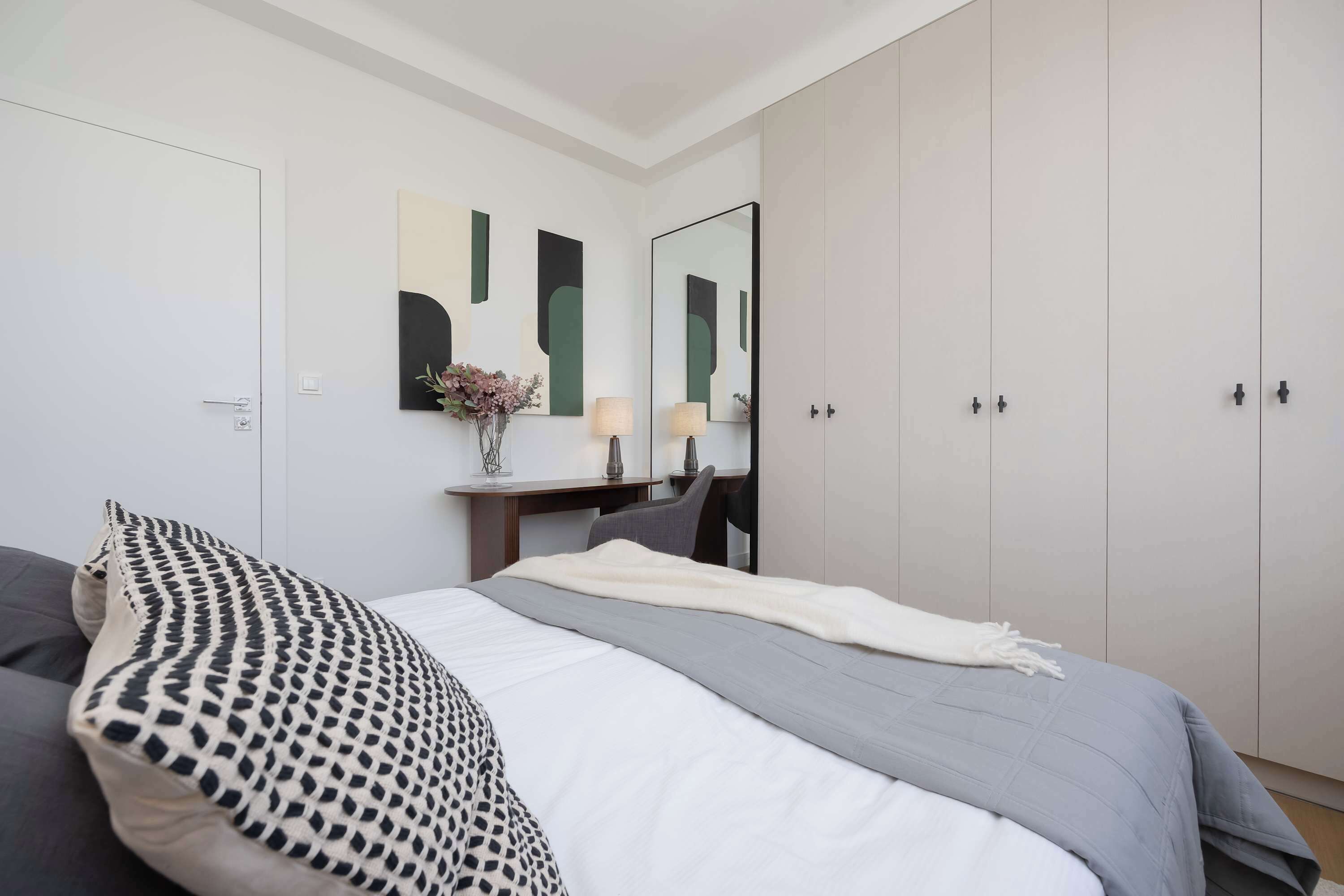 A bedroom with a bed, a desk, a chair, a wardrobe, and abstract wall art.