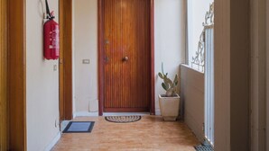 Un corridoio con una porta in legno, un estintore, una pianta in vaso e una finestra con decorazioni.