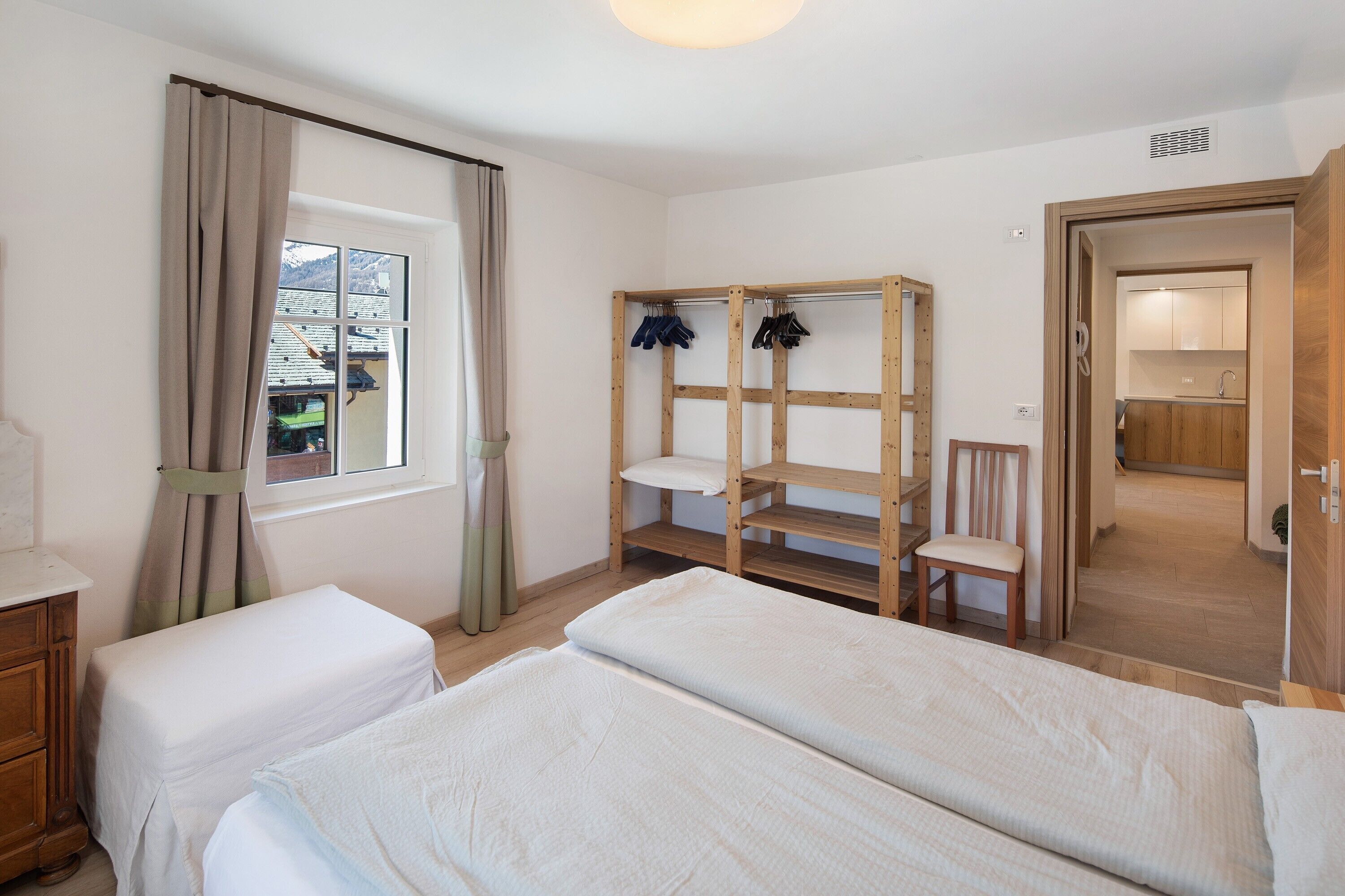 A bedroom with two beds, a wooden wardrobe, a chair, and a view of the outside.