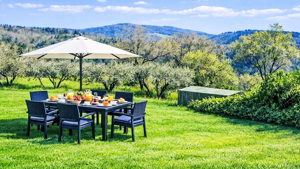 Ein Essbereich im Freien mit einem für das Frühstück gedeckten Tisch, umgeben von Stühlen und einem Sonnenschirm, mit Blick auf eine üppige Landschaft aus Bäumen und Hügeln.