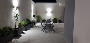 Uma área de refeições coberta ao ar livre, com mesa e cadeiras, rodeada por vasos com diversas plantas.
