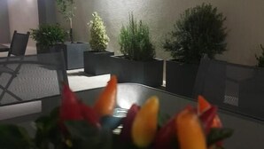 Un espacio al aire libre con mesas y sillas, rodeado de macetas con plantas y una lámpara fijada a la pared.