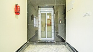 Un couloir étroit, avec un extincteur fixé au mur, une porte équipée d’une caméra de surveillance et un sol carrelé.