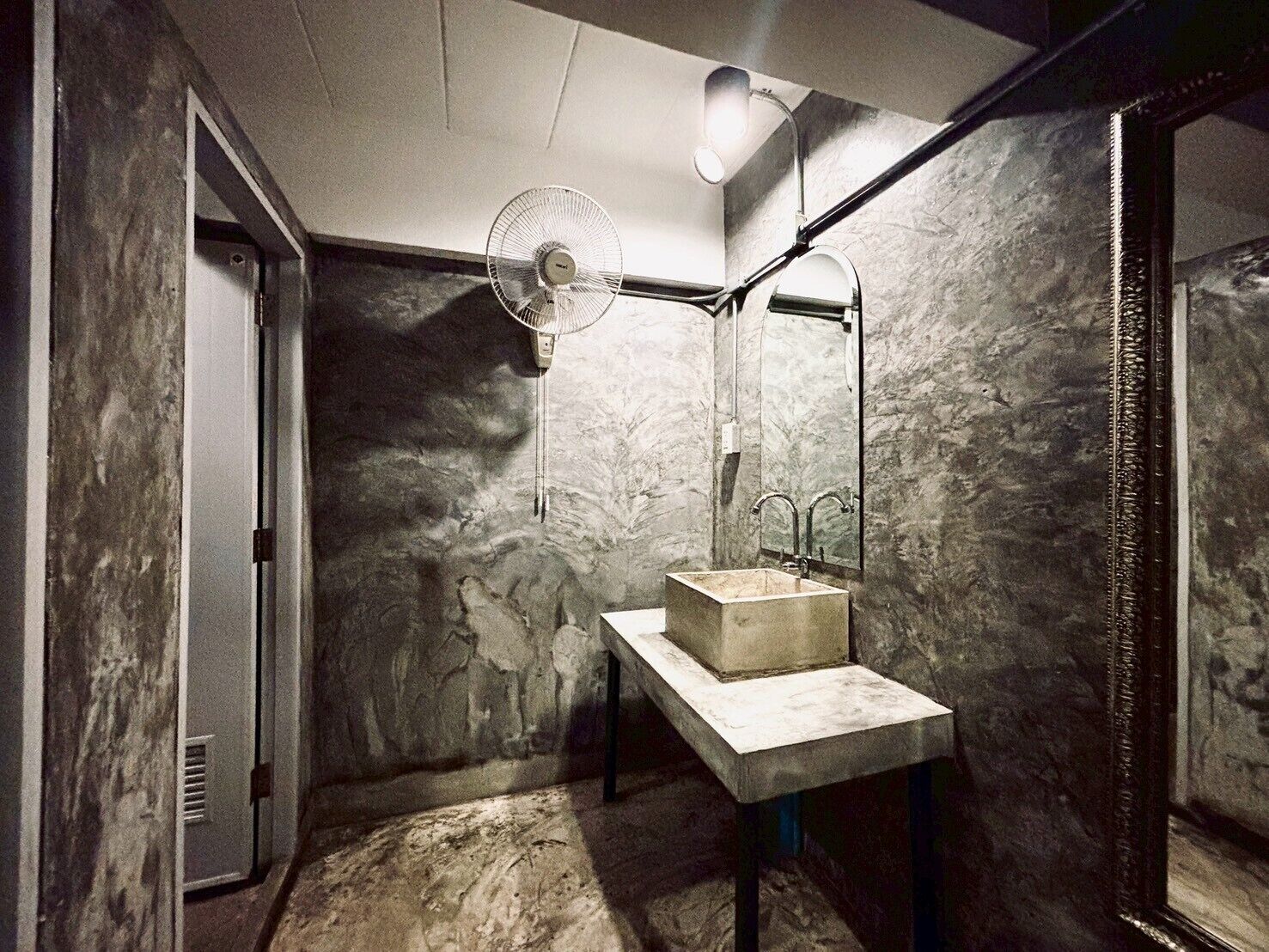 A bathroom with a marble wall, a pedestal sink, a mirror, a fan, and a light fixture.