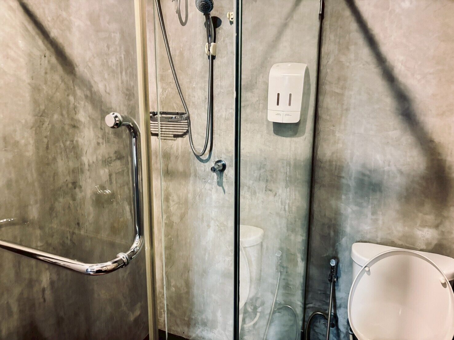 Un espace douche doté d’une porte en verre, d’une barre d’appui, d’un pommeau de douche à main, d’un robinet mural et de toilettes.