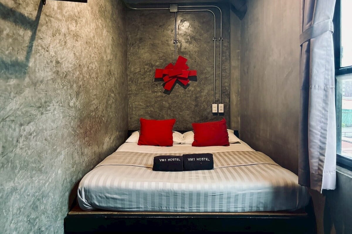 Une petite chambre comprenant un lit, deux oreillers rouges, un nœil rouge décoratif et deux étiquettes noires portant l’inscription « VM1 HOSTEL ».