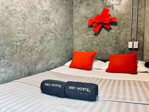 Une chambre d’hôtel avec un lit, deux oreillers rouges, une serviette noire avec l’inscription « VM1 HOSTEL », un nœud rouge décoratif et un mur gris texturé.