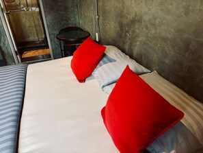 Uma cama com roupa de cama branca e almofadas vermelhas, uma mesa redonda e uma cadeira num quarto com parede de concreto.