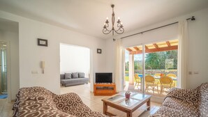 Ein Wohnzimmer mit gemusterten Sofas, einem Couchtisch, einem Fernseher und einer Glastür, die zu einem Außenbereich mit Pool und Stühlen führt.