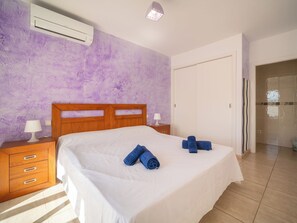 Une chambre à coucher avec une tête de lit en bois, un lit recouvert de draps blancs, deux serviettes enroulées, une table de chevet avec une lampe et un appareil de climatisation fixé au mur.