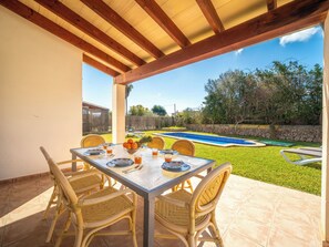 Une terrasse extérieure avec une table dressée pour quatre personnes, entourée de chaises en osier, donnant sur une piscine et une pelouse.