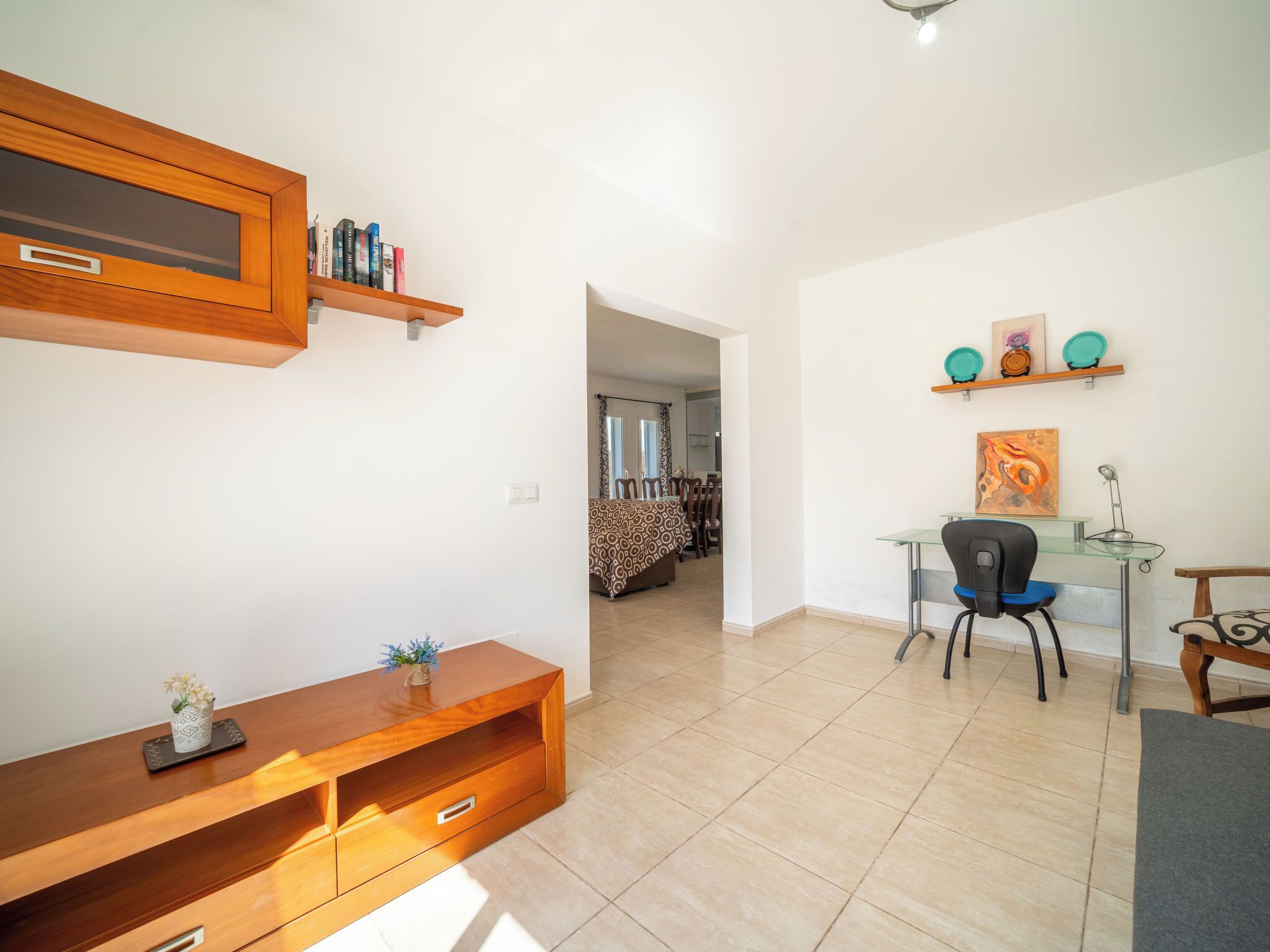 A modern living room with a wooden entertainment unit, a glass desk, and a dining area with a patterned sofa.