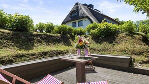 A patio with two striped lounge chairs, a table with a vase of flowers, and a house with a thatched roof in the background. - House Harvestehude apartment "Küstenglück" (Sylt)