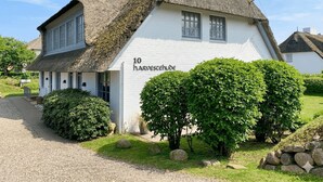 A white building with a thatched roof, labeled '10 harvesthuide,' surrounded by green bushes and rocks. - House Harvestehude apartment "Küstenglück" (Sylt)