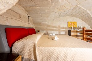 Una habitación con una cama, un almohadón rojo, una silla de madera y un cuadro en la pared.