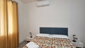 Un dormitori amb un llit, dues taules de nit, una petita taula auxiliar, una finestra amb cortines i un aparell d'aire condicionat.