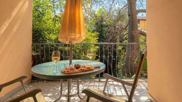 Une table ronde avec deux verres à vin, une assiette de nourriture et un parasol jaune sur un balcon doté d’une rambarde en métal et d’une végétation en arrière-plan.