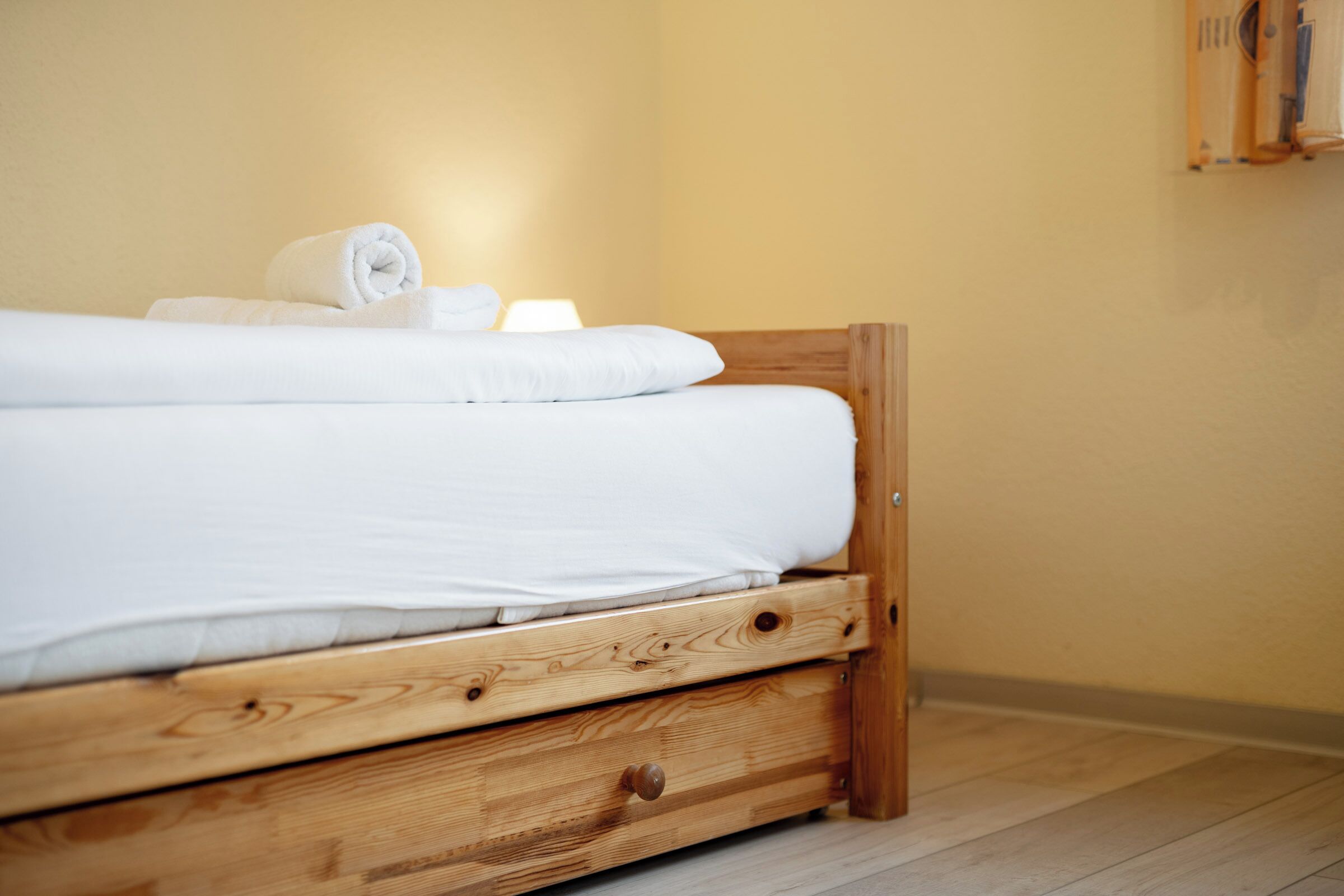 A wooden bed with white linens, a wooden drawer, and a wall-mounted towel rack.