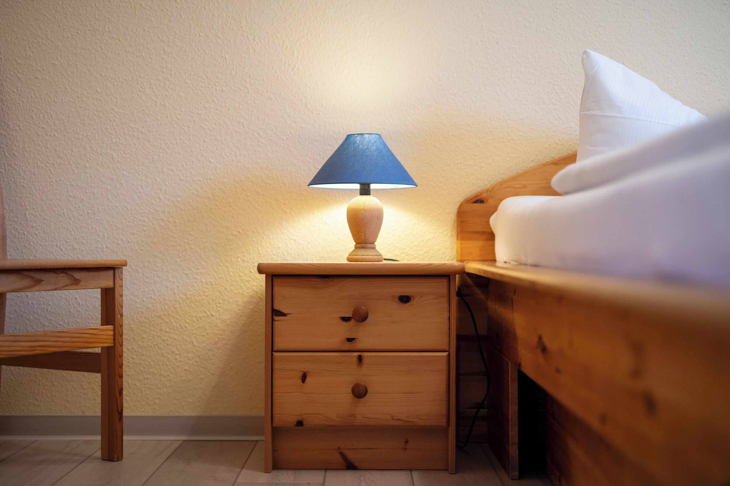 Una mesita de noche de madera con una lámpara, un cajón y una cama con ropa de cama blanca.