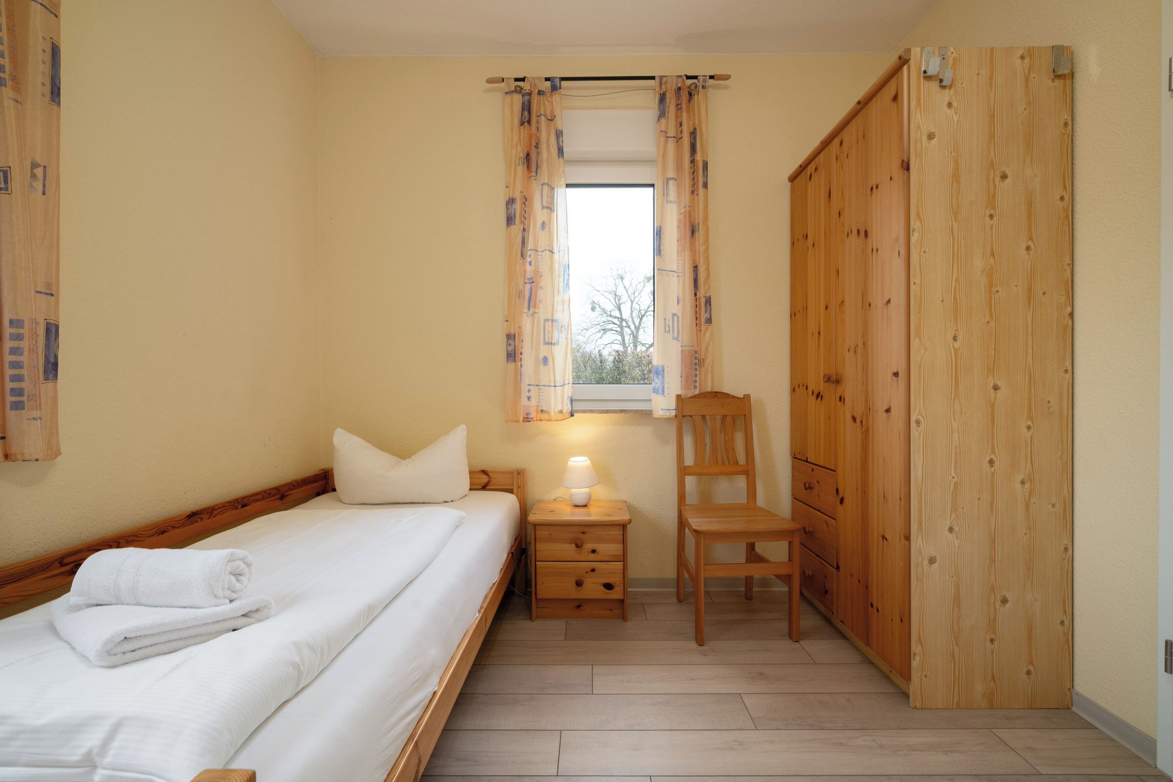 Une chambre à coucher avec un lit, une table de chevet avec une lampe, une chaise, une armoire et une fenêtre avec des rideaux.