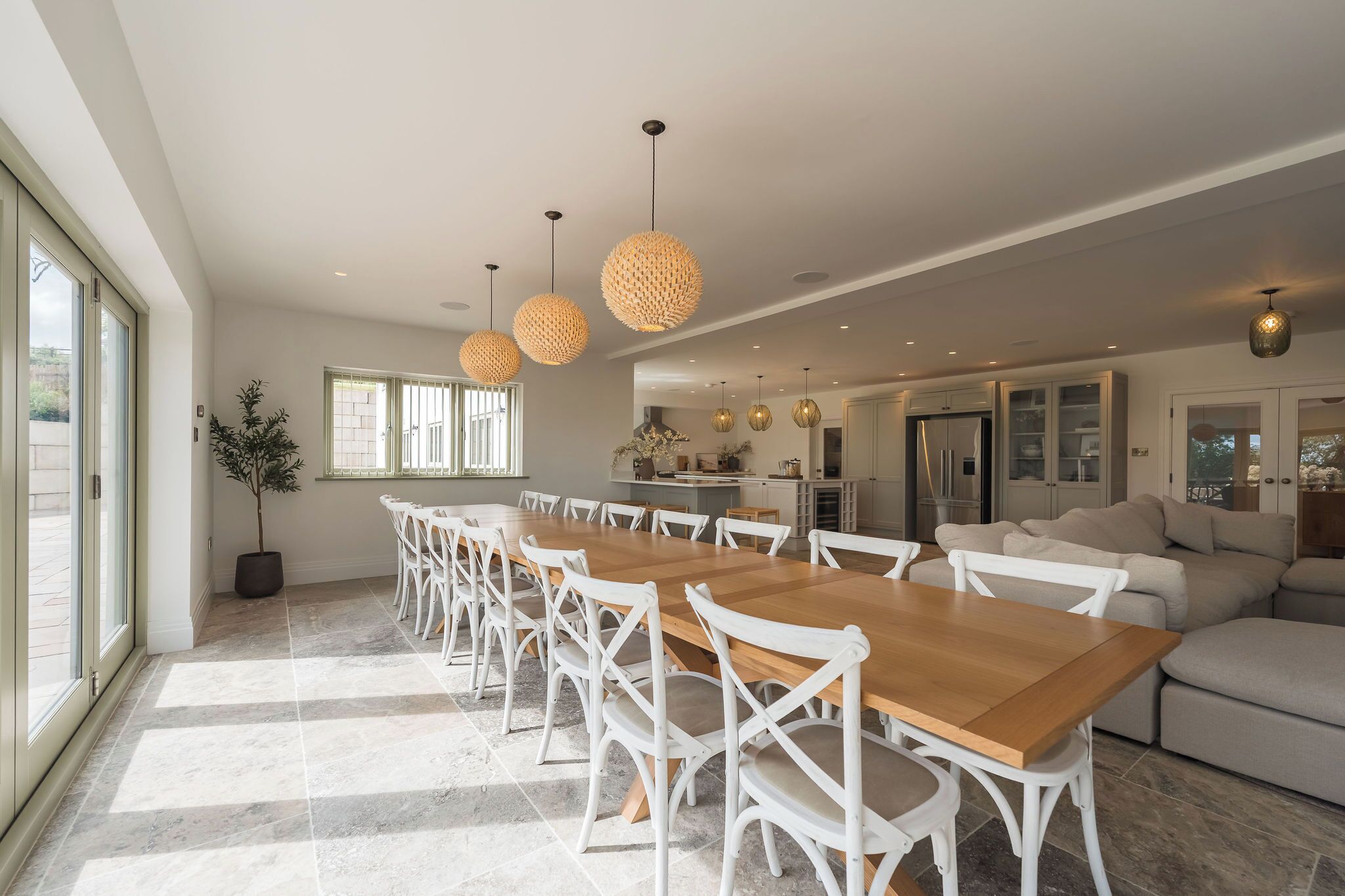 A spacious dining area with a long wooden table and white chairs, a kitchen in the background, and a large window providing natural light.