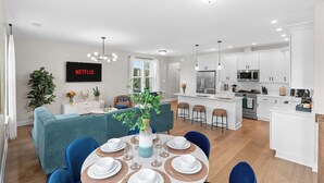 A modern kitchen with white cabinets, a central island, and a dining area with a blue sofa and round table. - Spacious 4BR w/ Basement Game Room • Lux Listings (Alexandria)