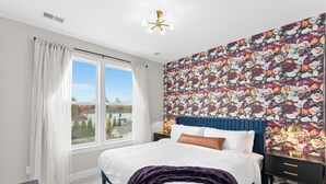 A bedroom with a large bed, a window with curtains, a floral wallpaper, a nightstand, and a ceiling light. - Spacious 4BR w/ Basement Game Room • Lux Listings (Alexandria)