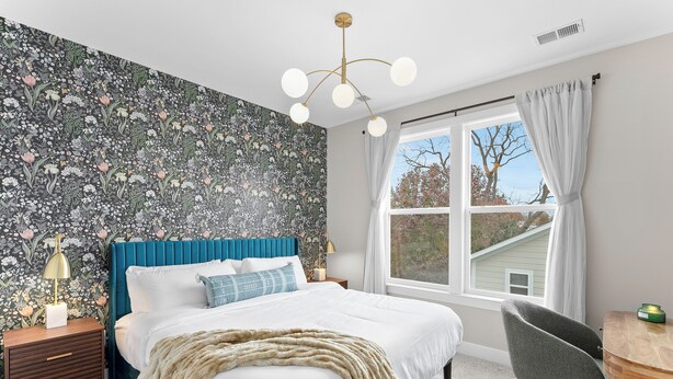 A bedroom with a floral wallpaper, a blue headboard, a white bed, a wooden nightstand, a lamp, and a view of the outside through a window. - Spacious 4BR w/ Basement Game Room • Lux Listings (Alexandria)