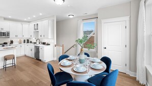 A modern kitchen with white cabinets, a dining area with blue chairs, and a view of a brick building through a window. - Spacious 4BR w/ Basement Game Room • Lux Listings (Alexandria)