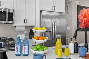 Una cocina con armarios blancos, electrodomésticos de acero inoxidable, un cuenco de frutas, botellas de agua y un exprimidor.