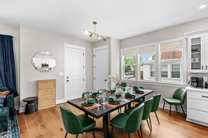 Eine moderne Küche mit weißen Schränken, ein Essbereich mit einem dunklen Tisch und grünen Stühlen sowie ein Fenster mit Blick auf ein Backsteinhaus.