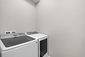 Ett tvättrum med en tvättmaskin och torktumlare från Samsung, en liten hylla med diverse föremål och en enkel vägg.