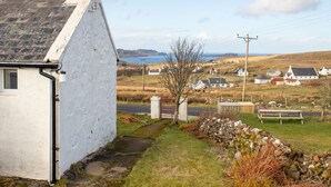 A white building with a chimney, a stone wall, and a bench in a rural setting with houses and a body of water in the background. - Ben Edra Croft by Interhome (North Skye)