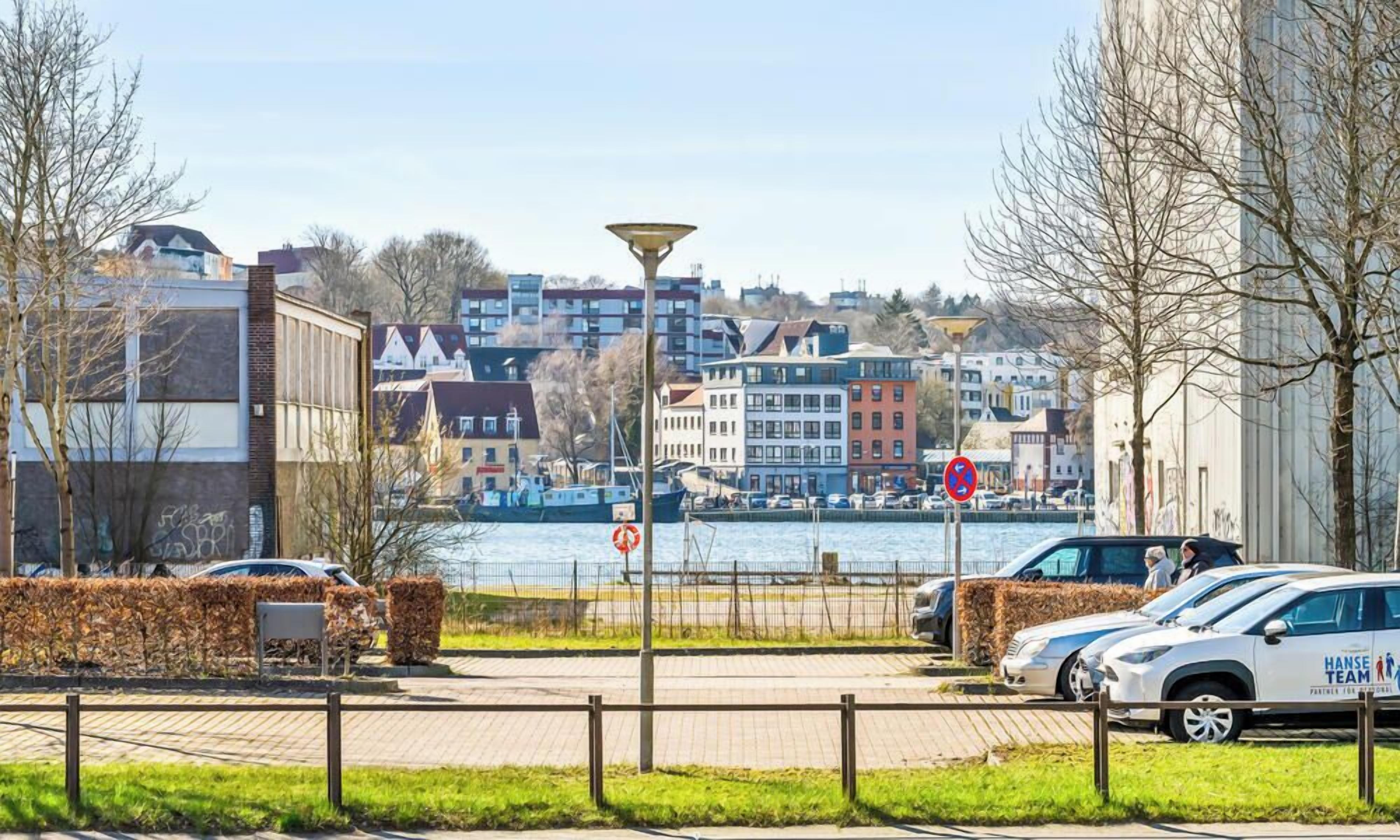Zona à beira-mar com carros estacionados, um caminho para pedestres e edifícios ao fundo.