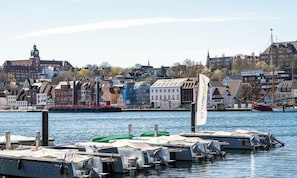 Um porto com barcos atracados, um edifício imponente com torre de relógio e um mastro com uma bandeira.
