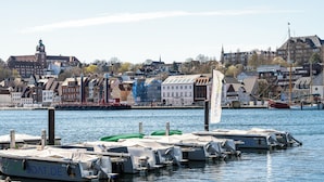 A harbor with boats docked, a prominent building with a clock tower, and a flagpole with a flag. - Steuerbord - own parking space, in the city, roller shutters, ground floor (Flensburg)