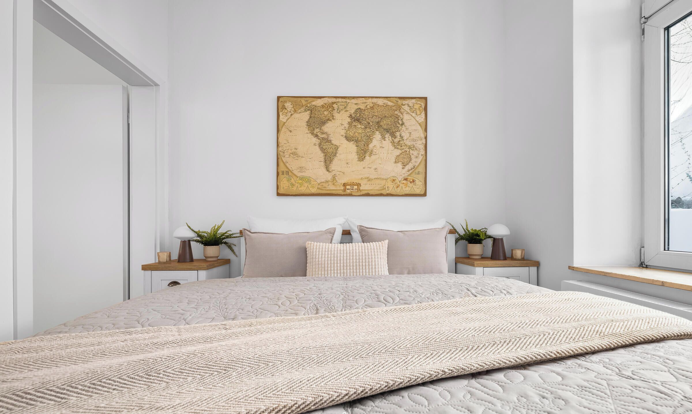 A bedroom with a large bed, a world map on the wall, bedside tables with lamps and plants, and a window with a view.