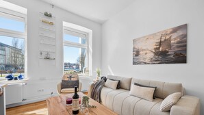A modern living room with a sofa, a wooden coffee table with a bottle of wine, and a painting of a ship on the wall. - Steuerbord - own parking space, in the city, roller shutters, ground floor (Flensburg)