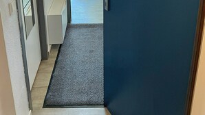 Ein Hotelzimmer-Eingang mit einer dunkelblauen Tür, einem kleinen Tisch mit einer Flasche, einem Stuhl und einem roten Handtuch, das an einem Haken hängt.