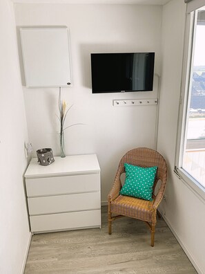 A room with a white dresser, a wicker chair with a polka dot cushion, a flat-screen TV mounted on the wall, and a window with a view. - Cozy Apartment with Stunning Views in Marina Wendtorf (Wendtorf)