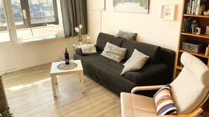 Ein Wohnzimmer mit einem dunkelgrauen Sofa, einem Holztisch, einem Bücherregal und einem Fenster mit Blick auf Gebäude.